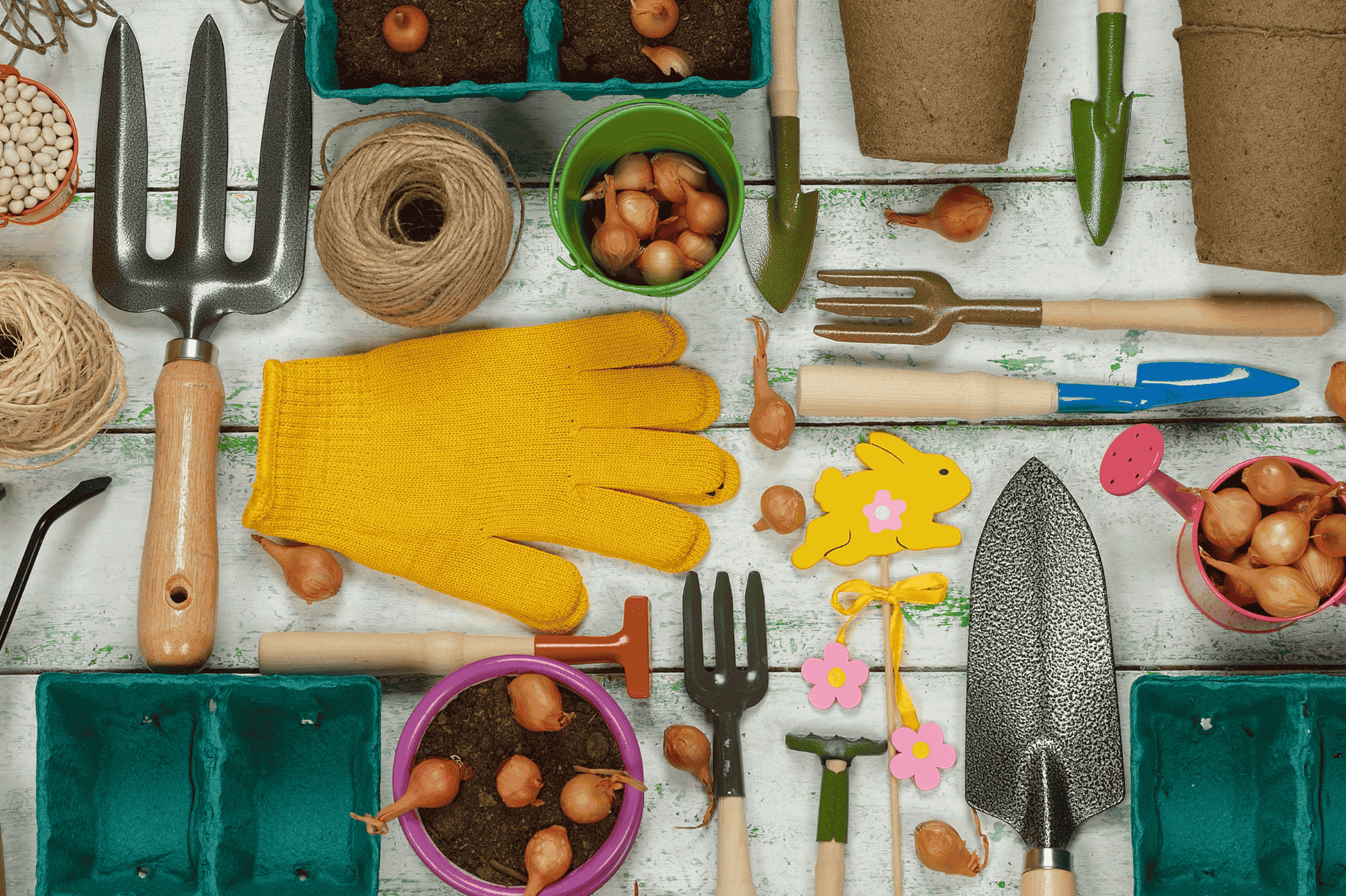 Gardening tools and supplies arranged on a wooden table, including pots, gloves, twine, and soil with green leaves nearby